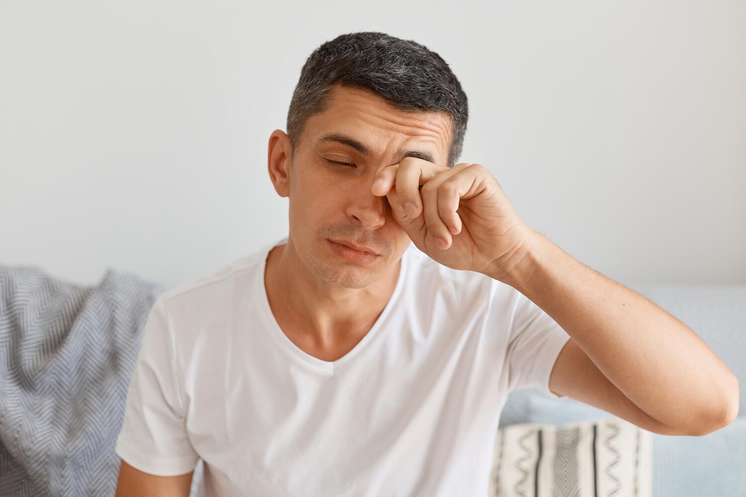 Tired exhausted young adult male with dark hair wearing white t shirt sitting comfortable sofa light living room rubbing his eyes has lack sleep feels eyes hurt 176532 16650